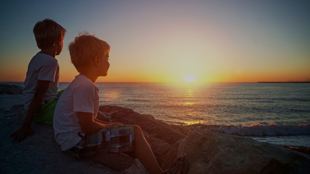 Two children stand on a beach, admiring the vibrant sunset over the Tuscan coastline.